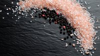 Pink Himalayan and black peppercorn salt crystals scattered in curved pattern on dark slate stone background, creating dynamic culinary composition.