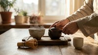 Traditional tea ceremony with hands pouring from ceramic teapot into cups on wooden tray, warm natural lighting creating peaceful mindful moment for wellness.