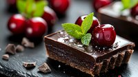 Chocolate tart garnished with fresh cherries, mint leaves and chocolate shavings on dark slate background, selective focus on dessert with blurred fruits.