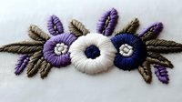 Delicate floral embroidery design featuring white, purple and navy blue flowers with olive leaves on white fabric, showcasing traditional needlework technique.