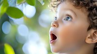 Young child with curly hair and open mouth expressing wonder and amazement while looking up at natural bokeh background with green leaves and sunlight.