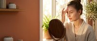 Woman with dark hair examining her face in mirror near window with terracotta wall and plants, skincare routine.