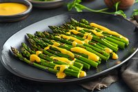Fresh green asparagus spears arranged on black plate drizzled with creamy hollandaise sauce, served on dark background for gourmet dining presentation.