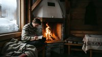 Young man in cozy sweater reading book by crackling fireplace in rustic cabin during winter, creating warm atmosphere for relaxation and comfort.