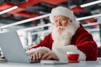 Santa Claus working on laptop computer in modern office during Christmas season. Holiday business concept with festive character typing at desk.