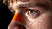 Close-up macro shot of blue eye with orange and red face paint or powder on nose and cheeks, showing freckles and skin texture in dramatic lighting.