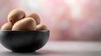 Fresh brown kiwi fruits in dark ceramic bowl on white surface with soft pink bokeh background for healthy eating and nutrition concepts.