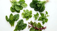 Fresh green leafy vegetables and herbs arranged on white background, including spinach, lettuce, and basil for healthy cooking and nutrition.