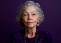 Elegant senior woman with silver hair wearing purple floral dress and statement necklace against dark background, expressing confidence and grace in studio portrait.