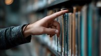 Female hand reaching for book on library shelf, wearing gray sweater sleeve, with blurred bookshelves background creating atmospheric depth and bokeh lighting effect.