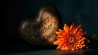 Wooden heart with Happy Mother's Day text beside vibrant orange chrysanthemum flower on dark background for greeting cards and holiday celebrations.