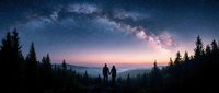 Silhouetted family standing together in forest landscape gazing at starry night sky with milky way galaxy and pink sunset horizon over mountains.