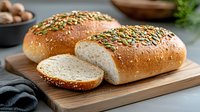 Fresh baked whole grain bread loaves topped with pumpkin seeds on wooden cutting board, one sliced, showing soft texture inside. Food photography.