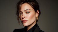 Elegant woman with glowing skin wearing black outfit and gold earrings against dark background, showcasing natural makeup and styled hair.