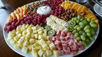 Fresh fruit, cheese and meat platter with dip arranged in colorful sections on white plate. Appetizing party food display includes raspberries, grapes, pineapple, nuts and cream sauce.