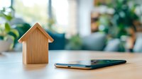 Wooden house model and smartphone on table against blurred green plants background, representing real estate app or smart home concept.