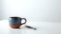 Handmade ceramic coffee mug with blue and terracotta glaze next to silver spoon on white table against neutral background for kitchen decor.