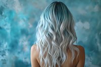 Young woman with long wavy platinum silver hair shown from behind against turquoise blue textured background, demonstrating modern hair coloring and styling technique.