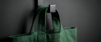Green canvas tote bag hanging on a black metal hook against a dark wall, eco-friendly reusable shopping bag in deep forest green color.