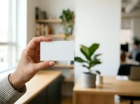 Hand holding blank white card in modern office workspace with natural light and potted plants, suitable for branding, business cards, and corporate marketing materials.