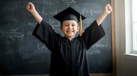 Happy young Caucasian boy in graduation cap and gown celebrating academic achievement with raised arms in classroom with mathematical formulas on blackboard.