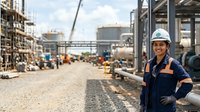 Female worker in hard hat and safety vest standing at industrial construction site with machinery and buildings in background, suitable for labor, engineering, and workplace safety concepts.