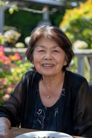 Senior Asian woman smiling warmly outdoors in garden setting, wearing elegant black outfit and silver necklace. Natural daylight illuminates genuine expression against floral background.