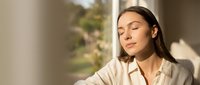Young woman with long dark hair and olive complexion wearing a cream-colored shirt, gazing thoughtfully by a window with natural sunlight. Suitable for wellness, lifestyle, and corporate projects.