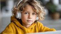 Young blonde curly-haired child wearing white headphones and yellow hoodie looking at camera in bright modern home interior for education and technology.