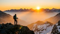 Hiker with backpack and trekking poles standing on mountain peak at sunrise, overlooking misty alpine valley and snow-capped peaks in golden morning light.