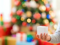 Cropped hand holding blank white gift box with festive decorated evergreen tree and colorful ornaments blurred in background for holiday season marketing and promotional campaigns.