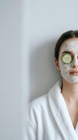 Young woman enjoying spa treatment with cucumber slice on eye and facial mask in white bathrobe for skincare and wellness routine at home.