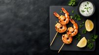 Grilled shrimp skewers served on black slate with creamy dipping sauce, lemon wedges, and fresh herbs against dark background.