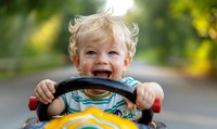 Cheerful toddler boy with blonde curly hair laughing while riding yellow toy car outdoors, showing pure joy and playful energy on summer day.