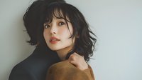 Young Asian woman with dark wavy hair and natural makeup wearing brown sweater poses against light wall, looking up with sensual expression. Professional portrait in soft lighting.