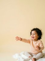 Happy toddler with dark curly hair and medium skin tone wearing peach sweater and white pants, sitting and reaching out with joyful expression against soft yellow background.