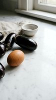 Fresh eggplants and brown egg arranged on marble counter with ceramic bowl in bright kitchen setting for cooking and meal preparation.