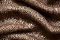 Natural brown linen fabric with visible weave texture creates soft waves and folds, showing detailed macro textile structure in warm sepia tones.