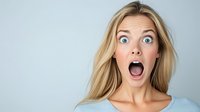 Young blonde woman with shocked surprised expression and wide open mouth against light blue background, wearing casual top. Emotional portrait.