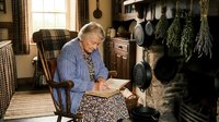 Elderly Caucasian woman in blue cardigan reading book by fireplace in cozy rustic kitchen with hanging herbs and cast iron cookware for homey lifestyle content.