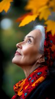 Senior woman with silver hair wearing vibrant red scarf gazes upward at golden autumn maple leaves against blurred natural background, expressing joy and serenity.