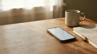 Smartphone and ceramic mug on wooden desk with notebook and pen, warm sunlight streaming through windows, workspace setup for productivity and creativity.