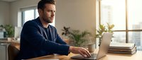 Focused Caucasian businessman working on laptop at wooden desk in bright modern office with plants and natural sunlight streaming through window.