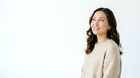 Young woman with long dark wavy hair and olive skin tone wearing a beige knit sweater, looking upward with a thoughtful expression against a white background.