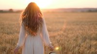 Young woman with long wavy hair in white dress standing in golden wheat field at sunset, back view, peaceful countryside scene for wellness concepts.