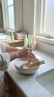 Woman hands applying serum from dropper bottle into white ceramic bowl with bamboo tools for natural skincare routine in bright bathroom window.