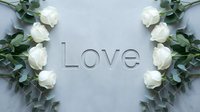 White roses and eucalyptus branches frame the word Love on a soft gray background, creating an elegant romantic flat lay for wedding or Valentine designs.