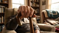 Senior man with weathered hands gripping wooden cane in cozy home library setting with bookshelves and armchair, representing aging, mobility assistance, retirement lifestyle.