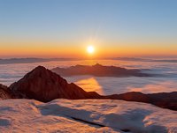 Majestic mountain peak rising above sea of clouds during golden sunrise, with snow-covered foreground and dramatic orange sky reflecting on misty valley below.