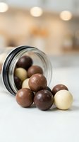 Mixed chocolate balls spilling from glass jar on kitchen counter with warm bokeh lights creating cozy atmosphere for confectionery and dessert concepts.